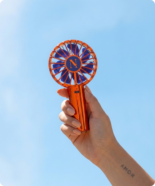 Een hand met de getatoeëerde tekst 'AMOR' houdt een oranje metallic Nimya draagbare ventilator omhoog tegen een helderblauwe lucht.