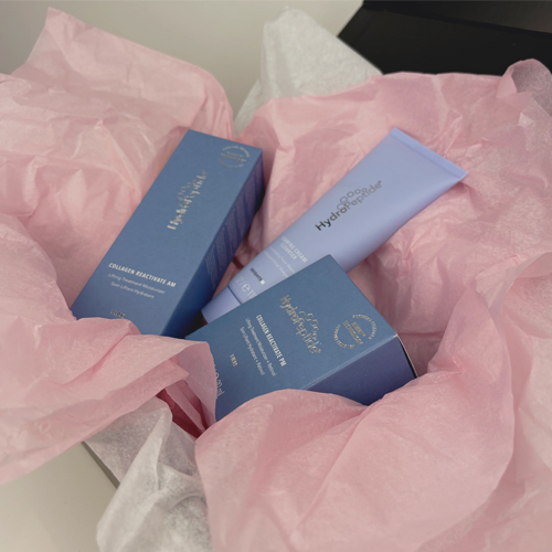 Close-up van de HydroPeptide Glow With Love producten, waaronder de Foaming Cream Cleanser en Collagen ReActivate AM, liggend in roze vloeipapier.