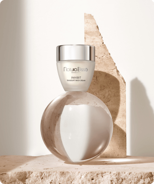 Een pot Natura Bissé Inhibit Tensolift Neck Cream balancerend op een glazen bol, geplaatst tegen een stenen achtergrond met zacht licht.