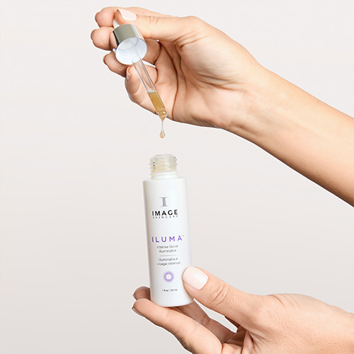 Een model houdt een geopende flacon Image Skincare Iluma Intense Facial Illuminator vast terwijl er een druppel serum uit de pipet valt.