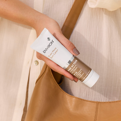 Een sfeerbeeld van een persoon die een tube Environ RAD Active Antioxidant Sun Cream SPF 20 vasthoudt boven een bruine leren tas. De tube heeft een wit en goudbruin ontwerp dat past bij de luxe uitstraling van het merk.