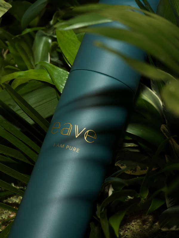 Een close-up van een donkergroene flacon van EAVE met de tekst "I AM PURE", omringd door groene bladeren in een natuurlijke setting.