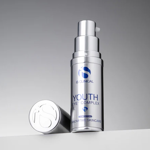 Een zilverkleurige flacon van de "iS CLINICAL Youth Eye Complex" oogcrème met de bijbehorende dop ernaast, staand op een witte ondergrond tegen een grijze achtergrond.
