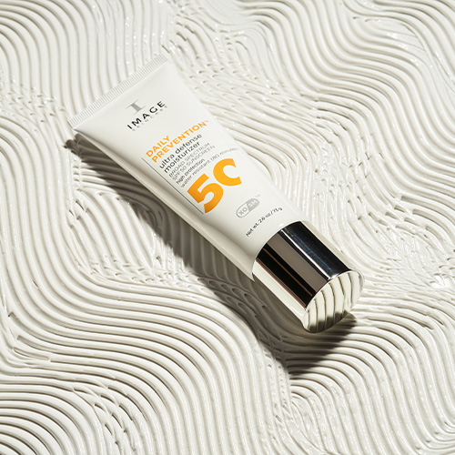 De Ultra Defense SPF 50 tube gepresenteerd op een artistiek uitgesmeerde ondergrond van witte zonnebrandcrème.