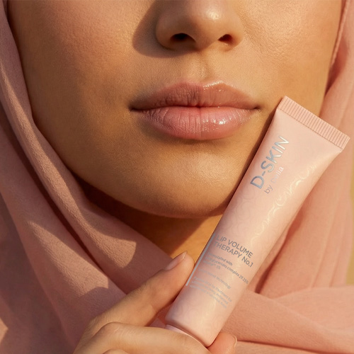 Een model houdt de roze tube van D-SKIN Lip Volume Therapy No. 1 vast bij haar gezicht. Ze draagt een bijpassende roze hoofddoek en heeft glanzende, gehydrateerde lippen in het warme zonlicht.