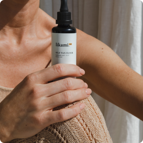 Vrouw met een zongebruinde huid houdt een glazen flesje Likami Self Tan Elixir vast voor haar schouder bij warm zonlicht.