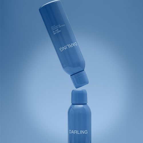 Twee blauwe flessen Darling Mist Me SPF 50 zonnebrand spray, waarvan één zwevend boven de andere, tegen een blauwe achtergrond met vignet.