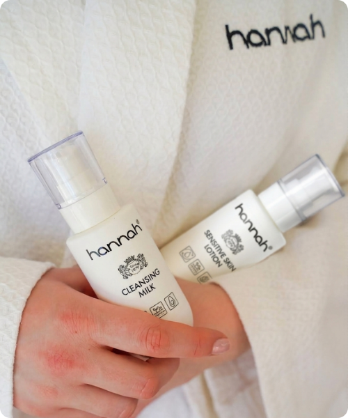 Sfeerbeeld van een persoon in een witte badjas met het hannah logo geborduurd op de kraag, die de hannah Cleansing Milk en Sensitive Skin Lotion in de handen draagt.