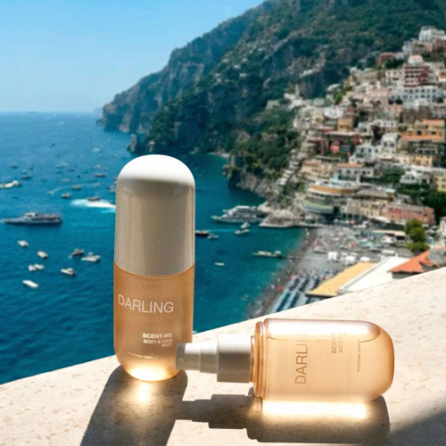 Twee flacons van de Darling Scent Me mist op een stenen balustrade met op de achtergrond een zonnig panoramisch uitzicht op de kust van Positano in Italie