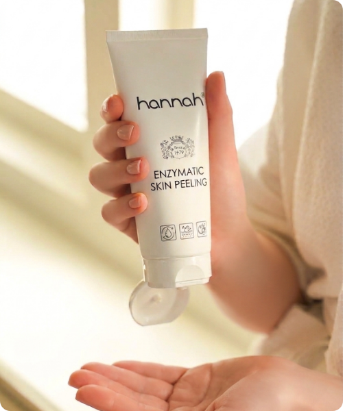 Een sfeerbeeld waarin iemand een witte tube hannah enzymatic skin peeling vasthoudt en het product in een open handpalm knijpt.