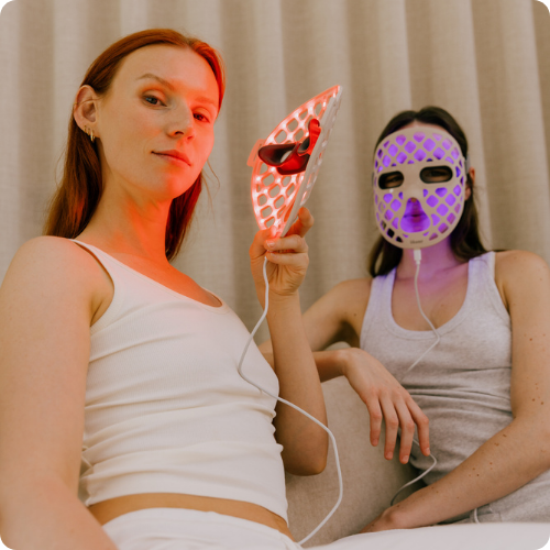 Twee vrouwen op een bank testen het Likami LED-masker met rood en paars licht. De afbeelding heeft afgeronde hoeken.