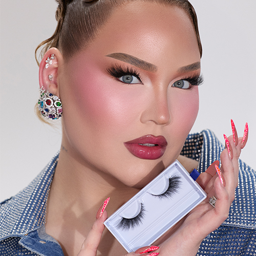 NikkieTutorials houdt de opengeschoven Nimya Lash It Real Good wimperverpakking vast in haar linkerhand. Ze draagt de wimpers, volledige make up, kleurrijke oorbellen en een denim jasje. Haar roze glitter nagels zijn zichtbaar.