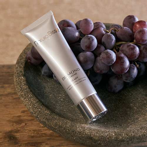 Een zilveren pot van het Natura Bissé Diamond Extreme Ice Lift masker, artistiek belicht met paarse tinten en een zachte reflectie op de ondergrond.