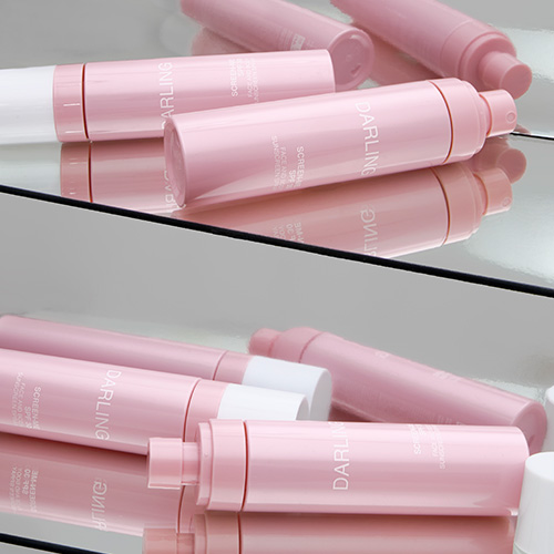 Verschillende roze flacons van de DARLING SCREEN-ME SPF 30 spray liggend op een spiegelend oppervlak.