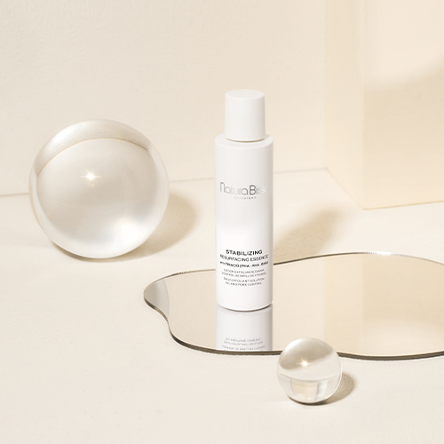De fles Natura Bissé Stabilizing Resurfacing Essence op een spiegeloppervlak, omringd door glazen bollen in een minimalistische setting.