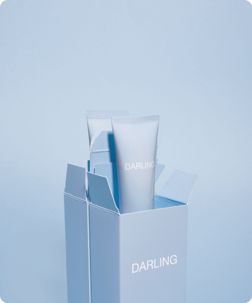 Een lichtblauwe tube van DARLING Glowy Screen die uit een geopende, bijpassende lichtblauwe kartonnen verpakking steekt tegen een egale blauwe achtergrond.