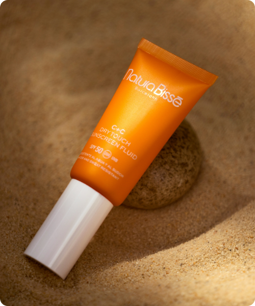 Een tube Natura Bissé C+C Dry Touch Sunscreen Fluid SPF50 liggend op het zand naast een steen.