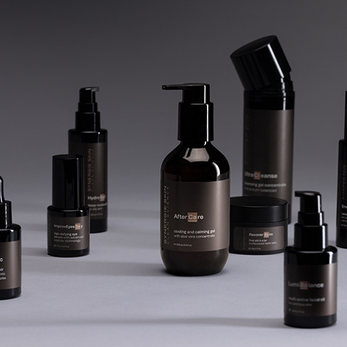 Een sfeerfoto van de Synergie Skin AfterCare fles in het midden omringd door verschillende andere cosmeticaproducten en serums van het merk tegen een egale grijze achtergrond.
