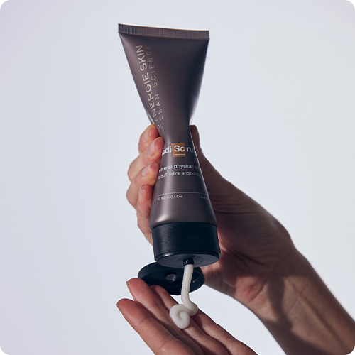 Handen die een donker grijze/bruine tube Synergie Skin MediScrub vasthouden en een hoeveelheid witte gezichtsscrub op de palm knijpen.