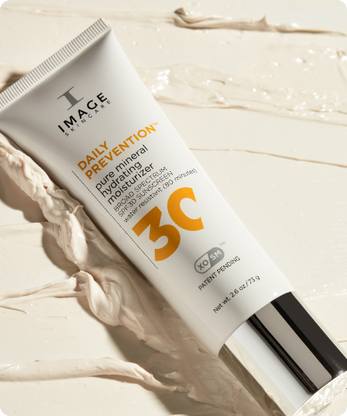 Een tube IMAGE Skincare Pure Mineral Hydrating Moisturizer met SPF 30, liggend op een lichte achtergrond van romige, wit-beige productstrepen. De tube heeft een zilveren dop en een groot gele getal "30" op de verpakking.