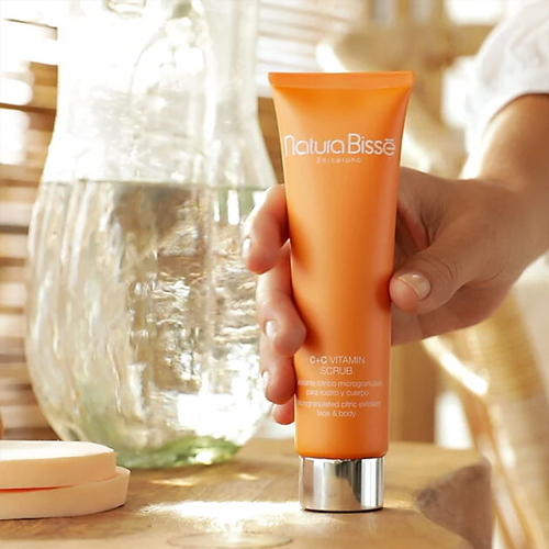 Iemand houdt de oranje tube C+C Vitamin Scrub vast in een lichte, zomerse omgeving met een glazen karaf op de achtergrond.