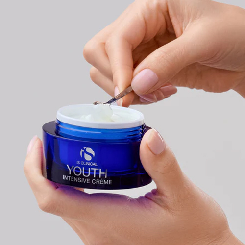 Handen die een blauw potje iS Clinical Youth Intensive Crème vasthouden en met een kleine cosmetische spatel wat van de witte crème eruit scheppen