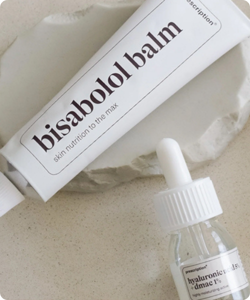 Een close-up sfeerbeeld van een witte tube Prescription Bisabolol Balm liggend op een stenen ondergrond, met op de voorgrond een glazen pipetflesje Prescription Hyaluronic Acid 5% + DMAE 1%