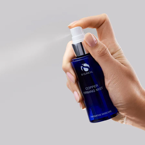 Hand die de donkerblauwe sprayflacon van iS Clinical Copper Firming Mist vasthoudt en een fijne, verfrissende nevel sprayt tegen een grijze achtergrond