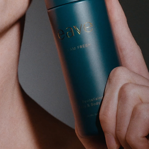 Close-up van een model dat een donkergroene flacon van Eave I AM FRESH vasthoudt bij de hals en schouder.