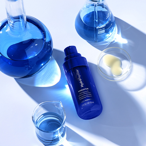 De blauwe fles HydroPeptide Hydrostem omringd door laboratoriumglaswerk met blauwe vloeistof en een petrischaaltje met serumdruppels.