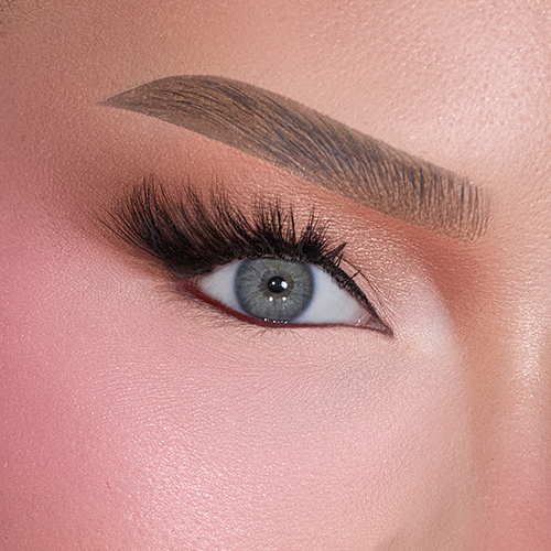 Een close up foto van één oog met grijze iris, gedefinieerde wenkbrauw, zwarte eyeliner en volle nepwimpers op een huid met roze blos.