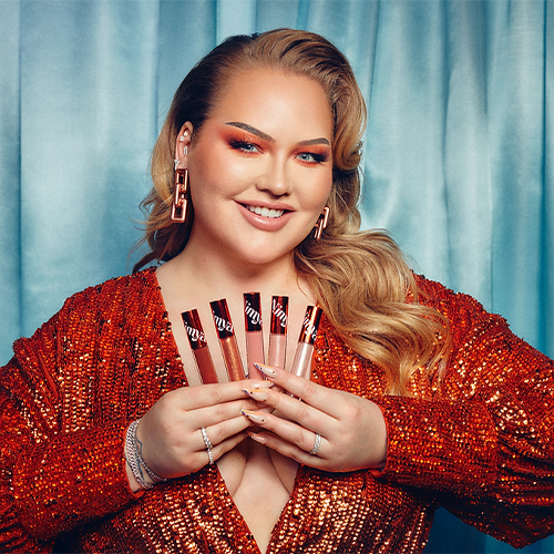 NikkieTutorials glimlachend in een rode top met in beide handen een selectie Nimya Spill The Juice lipglosses voor een blauw gordijn