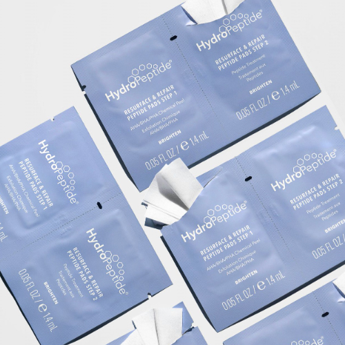 Meerdere geopende en gesloten blauwe sachets van de HydroPeptide Resurface & Repair Peptide Pads Step 1 en Step 2, verspreid over een witte achtergrond met de witte exfoliërende pads zichtbaar.