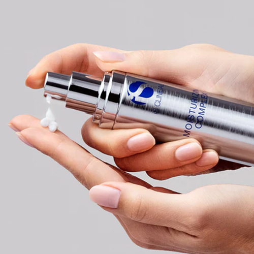 Handen die een witte, hydraterende crème pompen uit de zilveren flacon van iS Clinical Moisturizing Complex in de palm van de hand