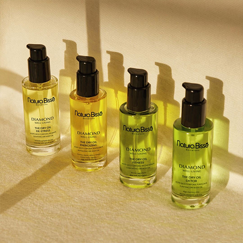 Vier flacons Natura Bissé body oil op een rij, werpend lange schaduwen in een warme, zonverlichte setting.