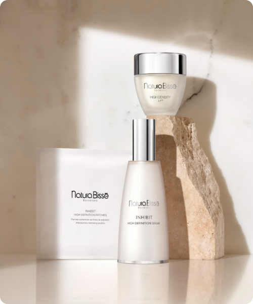 productfoto van de Natura Bissé Inhibit collectie, met het High Definition Serum en een verpakking High Definition Patches op de voorgrond, en het High Density Lift potje gebalanceerd op een ruwe steen tegen een marmeren achtergrond