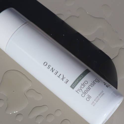 Sfeervolle productfoto van de fles Extenso Hydro Cleansing Oil (250ml) schuin liggend op een donkergrijs oppervlak, omgeven door waterdruppels.