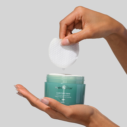 Hand van een model haalt een ronde HydroPeptide clarifying toner pad uit een groene verpakking