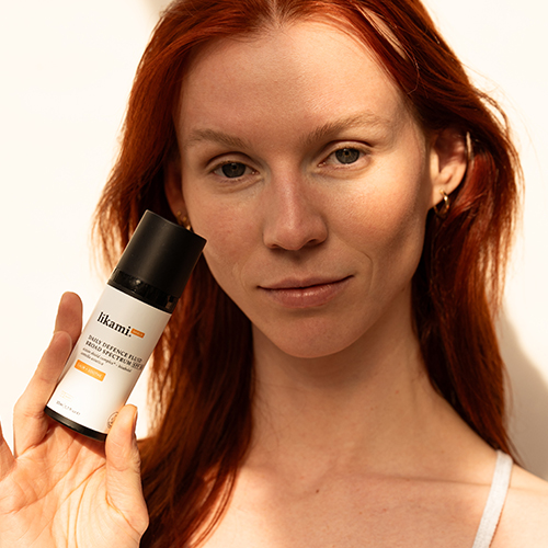 Een vrouw met rood haar houdt de Likami Daily Defence Fluid Calm + Soothe flacon dicht bij haar gezicht in warm, natuurlijk zonlicht.