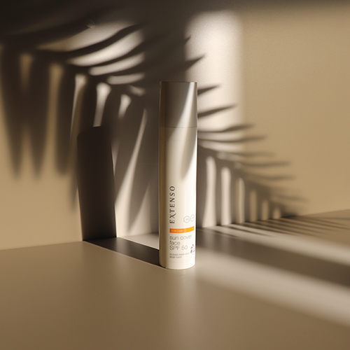 Een fles Extenso Sun Cover Face SPF 50 geplaatst in een sfeervolle setting, waarbij de schaduw van een palmblad over het product en de ondergrond valt.