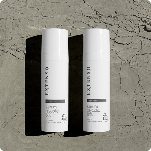Twee flacons van het Extenso Deep Peel serum glycolic 6% staan rechtop naast elkaar op een droge, gebarsten aarde-ondergrond in het felle zonlicht.