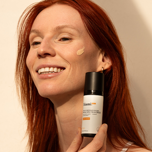 Close-up van een lachende vrouw met rood haar, met een kleine hoeveelheid van de getinte SPF-vloeistof op haar wang aangebracht.