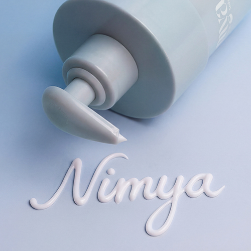 Close-up van de pomp van de Nimya bodylotion met de witte crème die het merknaam spelt op een blauwe ondergrond