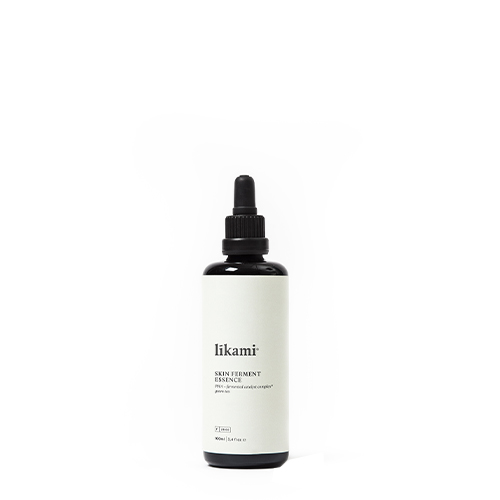 Een studiofoto van een enkele, donkere glazen fles met een zwarte druppeldop, gevuld met een transparante vloeistof. De fles staat op een witte achtergrond en heeft een crèmekleurig etiket met de tekst "likami SKIN FERMENT ESSENCE".