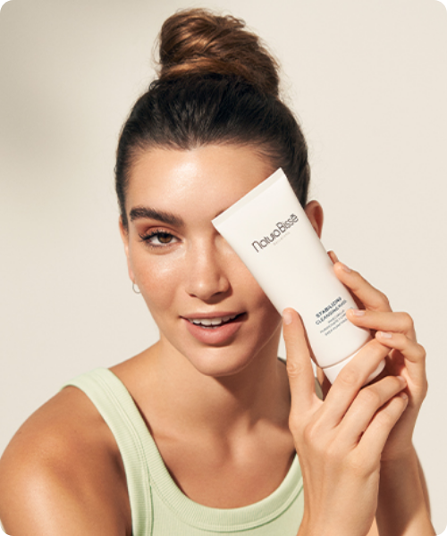 Een lachende vrouw met haar haar in een knot houdt een witte tube van het Natura Bissé Stabilizing Cleansing Mask voor haar gezicht tegen een lichte achtergrond.
