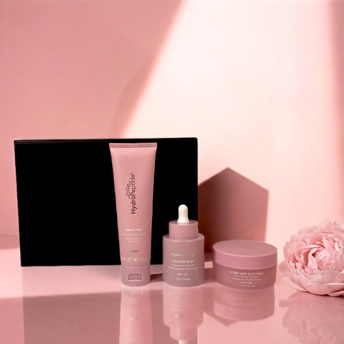 Drie roze HydroPeptide producten uit de Care With Love set, waaronder de Makeup Melt en Moisture Reset, sfeervol gefotografeerd met een roze bloem.