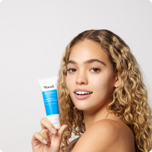 Een glimlachend model met krullend haar houdt een blauwe tube Murad Rapid Relief Acne Sulfur Mask vast