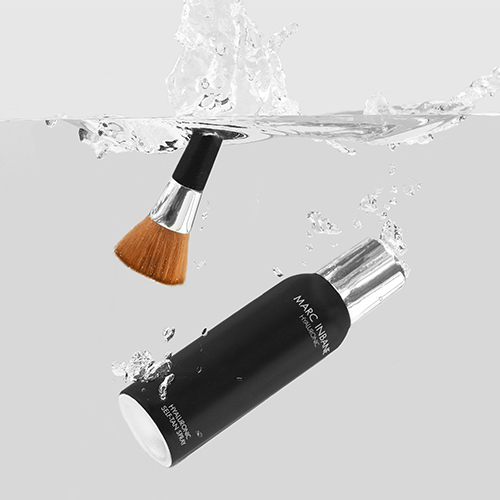 Sfeerafbeelding van de Marc Inbane Hyaluronic Self Tan Spray flacon en een tanning brush die ondergedompeld zijn in helder water met luchtbellen en opspattend water
