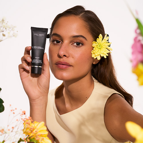 Een vrouw houdt de Marc Inbane Crème Bronze SPF30 tube vast te midden van bloemen, met een zonnige, egale teint.
