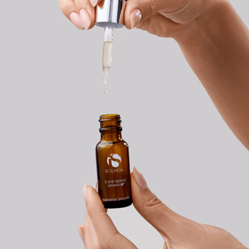 Iemand houdt een bruin glazen flesje iS CLINICAL C Eye Serum Advance+ vast en brengt met de druppelaar een druppel serum aan.